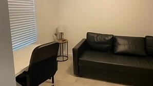 Ein Wohnzimmer mit einer schwarzen Ledersofa, einem schwarzen Sessel, einem kleinen Beistelltisch mit einer Lampe und einem Fenster mit Jalousien.