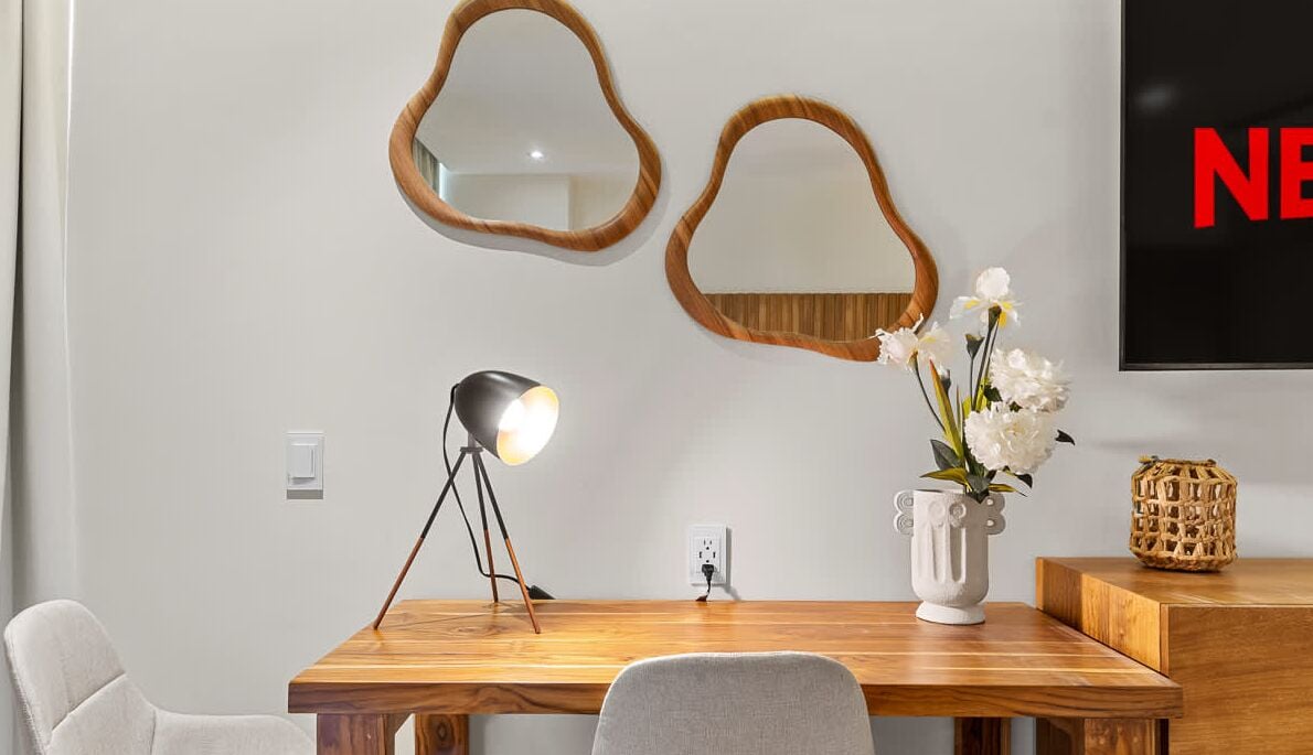 A modern room with a wooden desk, a lamp, a vase with flowers, and two wall mirrors.