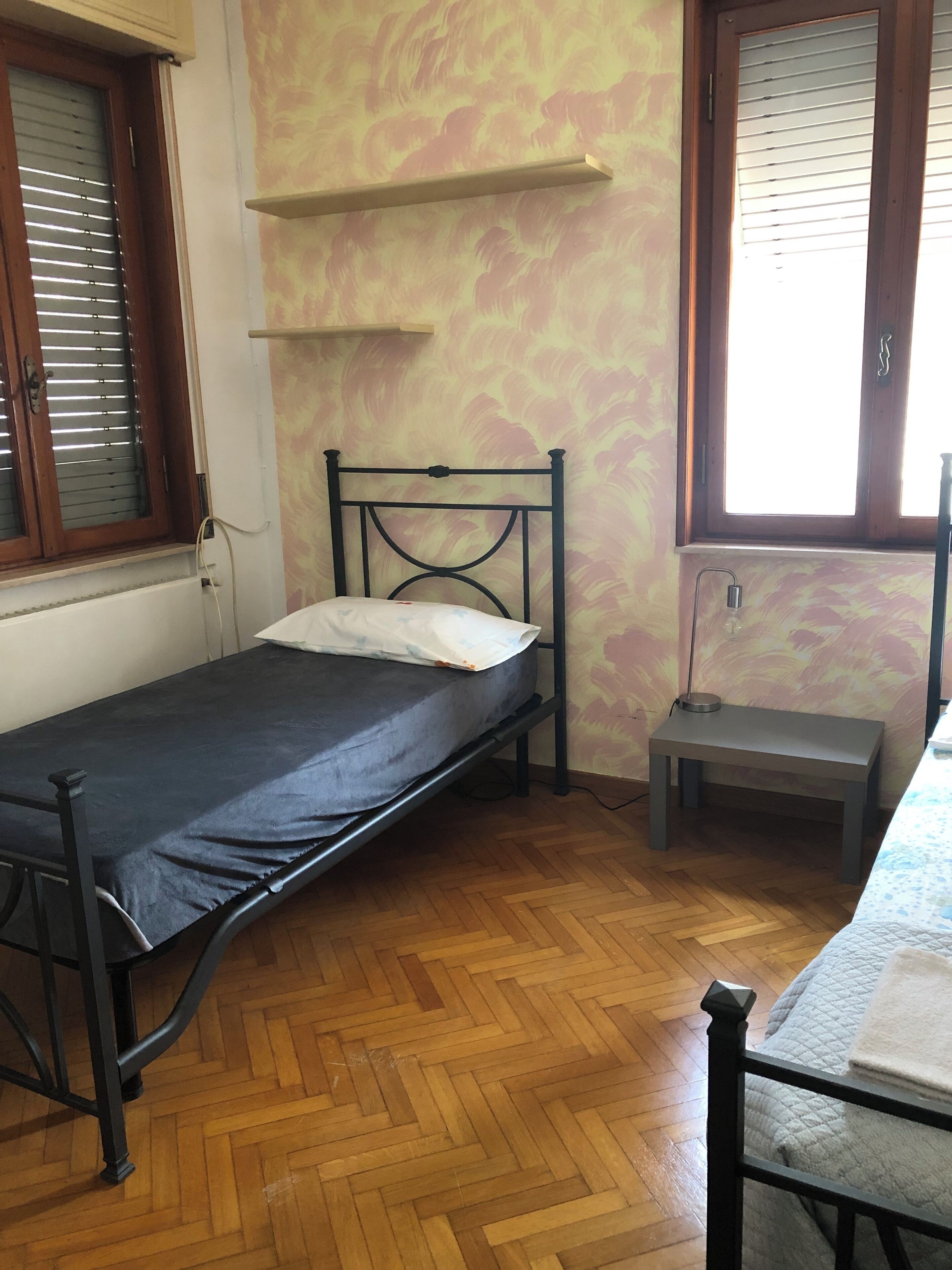 A single bed with a metal frame, a small bedside table, and a window with wooden blinds.