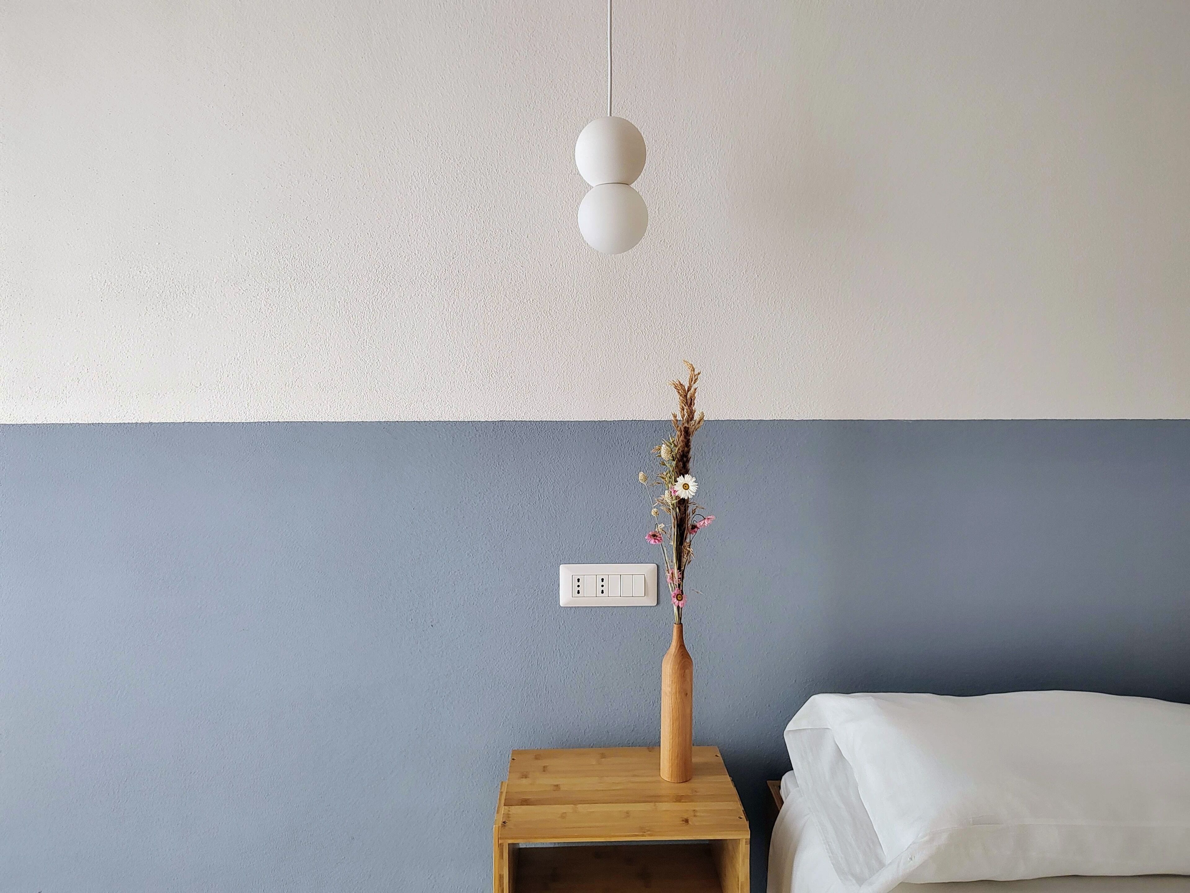 Quarto com mesa de cabeceira de madeira, um vaso com flores secas, cama branca e interruptor de parede.