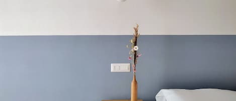 Quarto com mesa de cabeceira de madeira, um vaso com flores secas, cama branca e interruptor de parede.