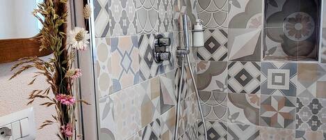 A bathroom with patterned tiles, a shower, a wooden vase with flowers, and a white sink.