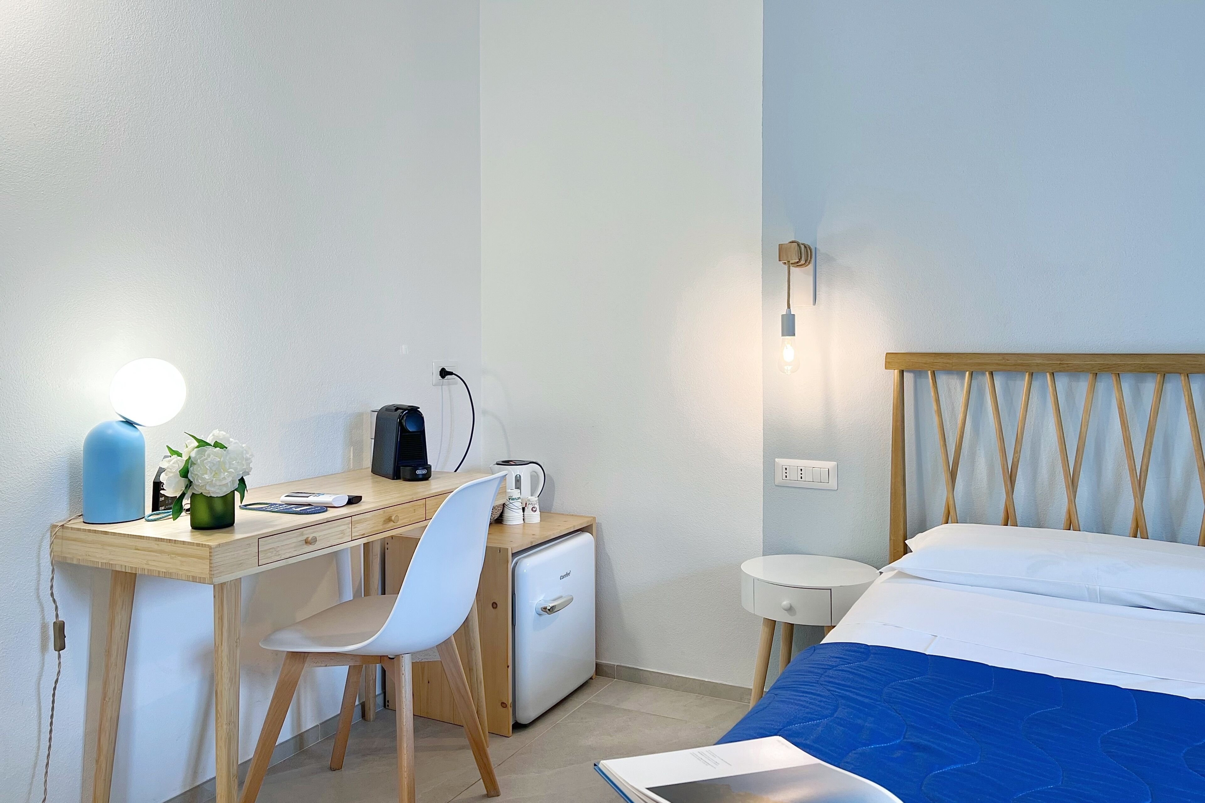 Een hotelkamer met een bed, een bureau met stoel, een kleine koelkast, een lamp en een wandlamp.