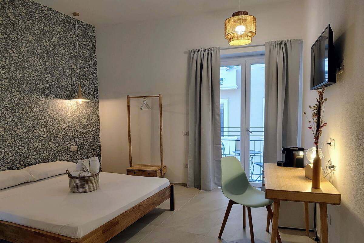 A bedroom with a bed, a desk, a chair, a hanging light fixture, a flat-screen TV, and a view of the outdoors through a window.