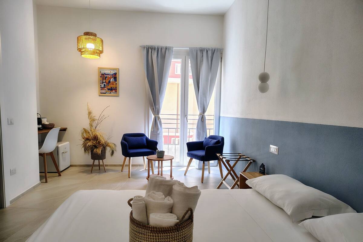 A hotel room with a bed, two chairs, a small table, a lamp, a picture on the wall, and a window with curtains.