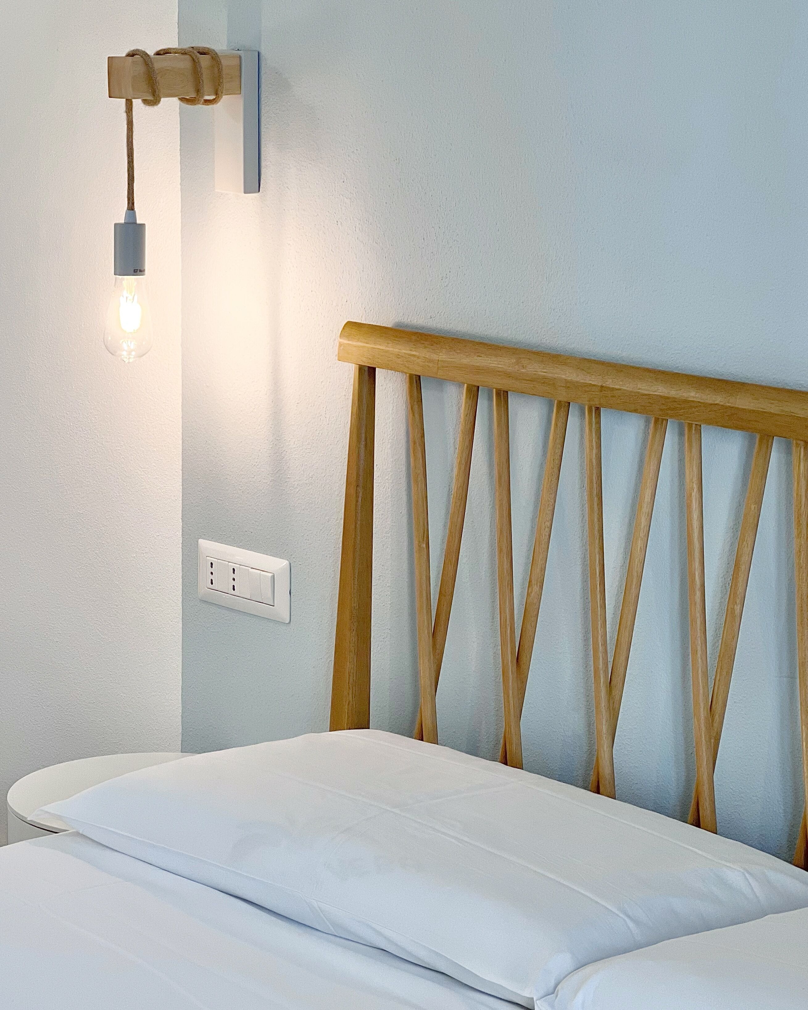 A bed with a wooden headboard, a wall-mounted light fixture, and a power outlet.