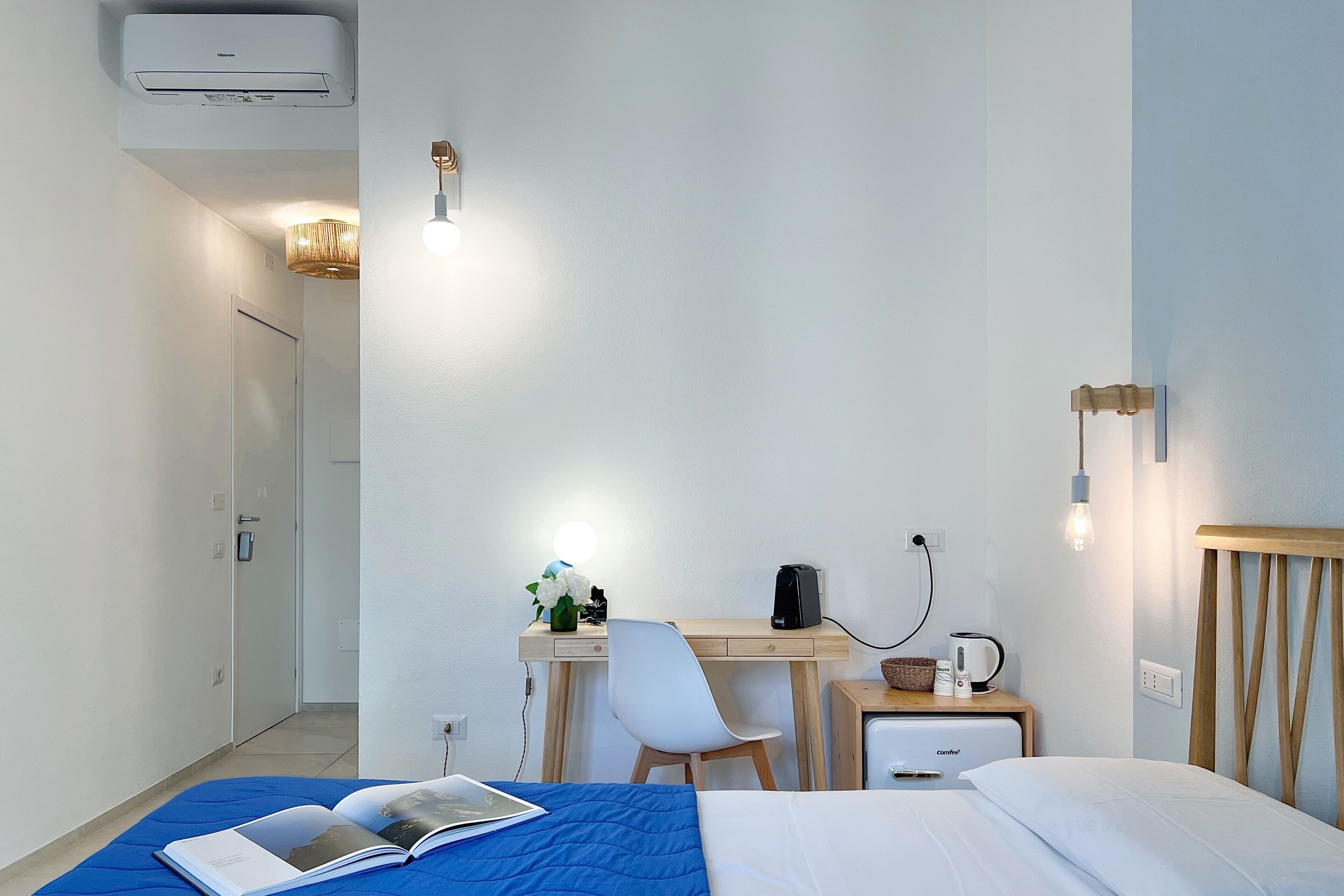 Een hotelkamer met een bed, een bureau met stoel, een nachtkastje met lamp en een airconditioning.