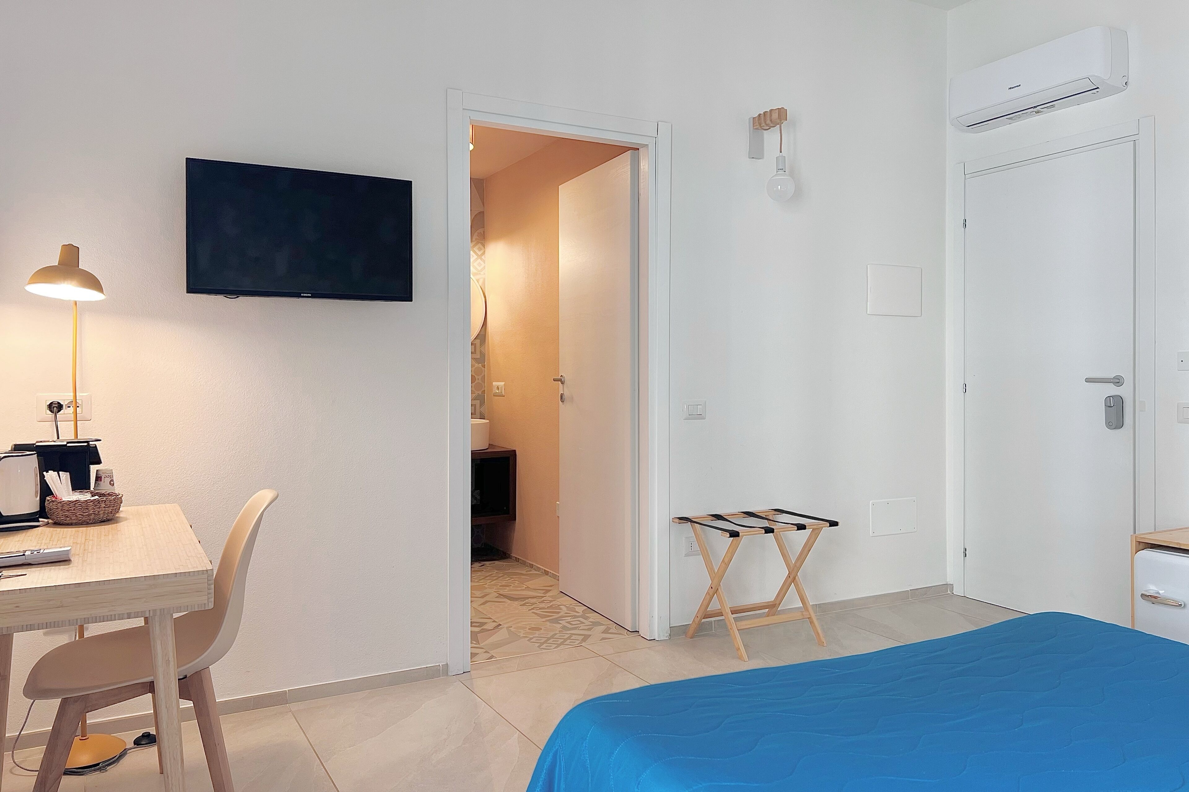 A hotel room with a bed, a desk with a lamp, a television, an air conditioning unit, and a door.