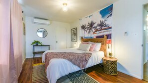 Un dormitorio con cama, mesita de noche, lámpara, espejo y pared decorada con un mural tropical.