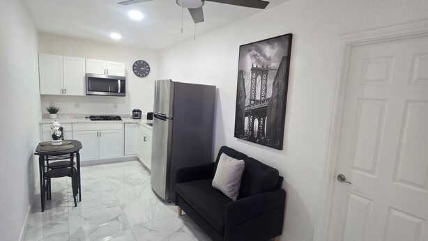 A compact living space with a kitchen, a black sofa with a cushion, a dining table with chairs, a ceiling fan, and a door.
