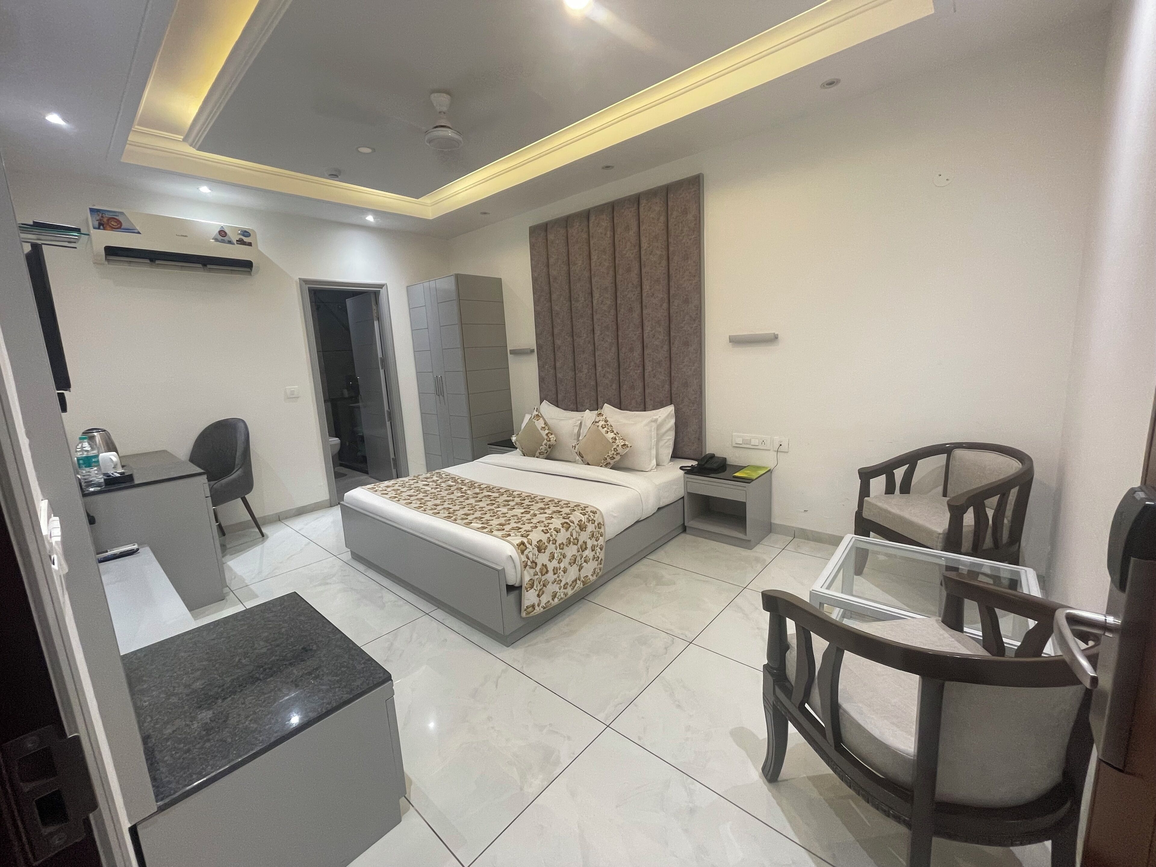 A modern hotel room with a bed, bedside table, desk, chairs, and an air conditioning unit.