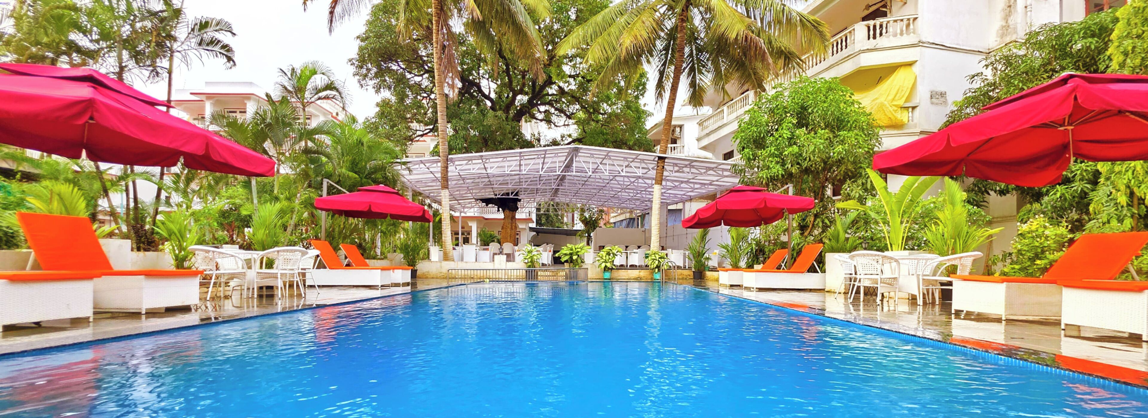 A swimming pool with red umbrellas, white lounge chairs, and a covered seating area surrounded by palm trees and greenery.