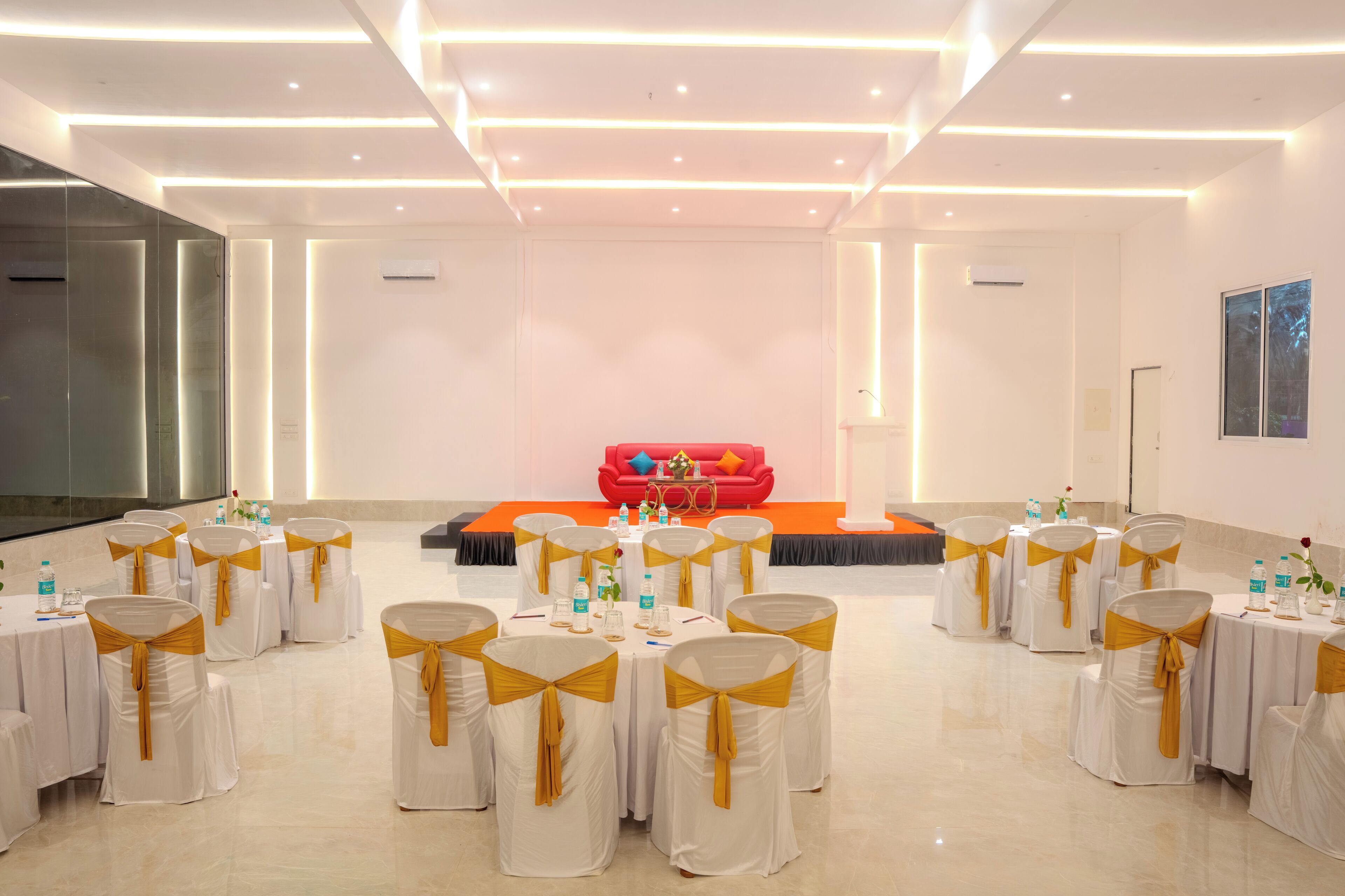 A conference room with white tables and chairs, a stage with a red sofa, and a modern ceiling design.