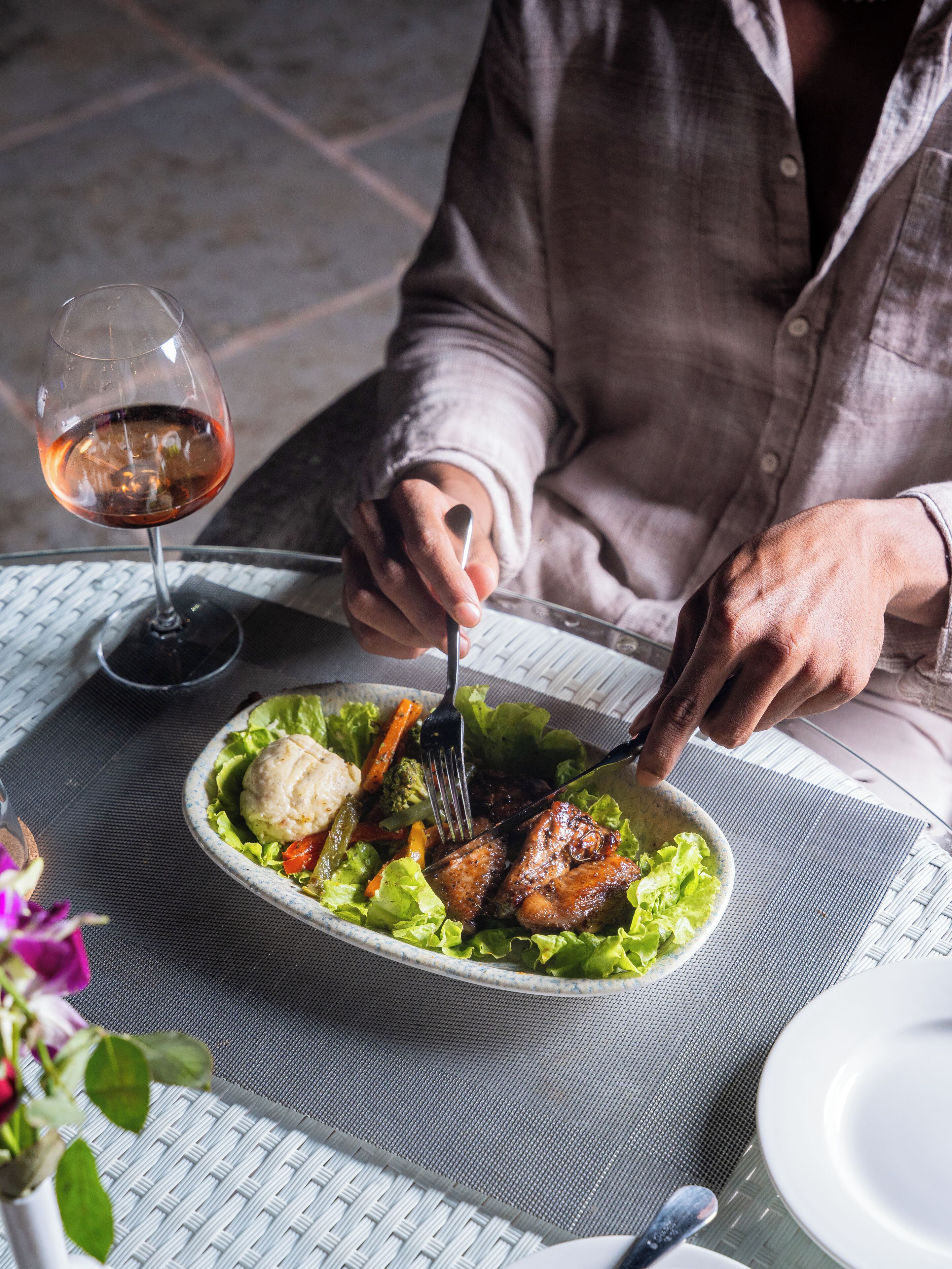 A person dining at an outdoor table with a meal, a glass of red wine, and a flower vase.