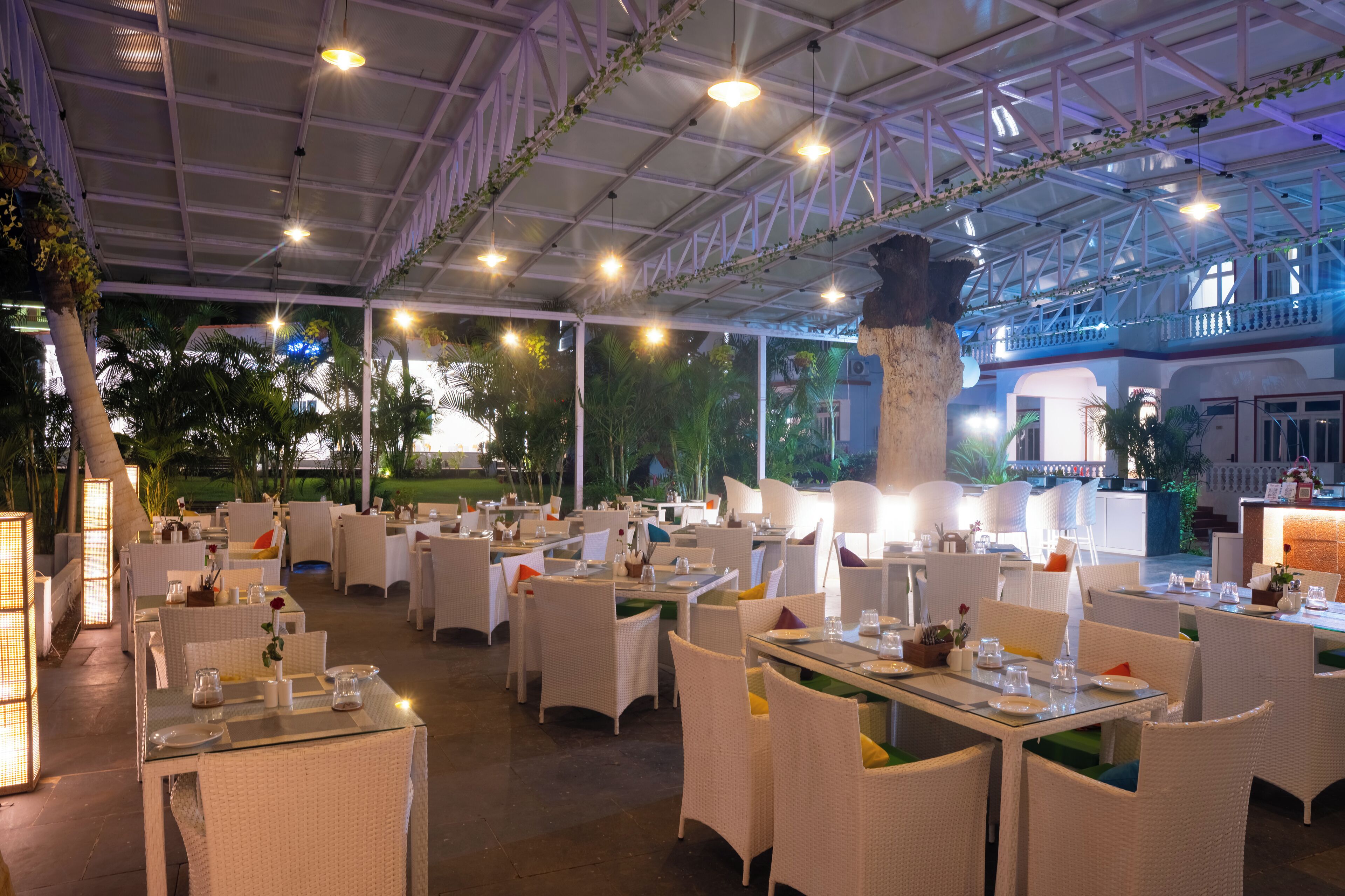 An outdoor dining area with white wicker furniture and glass tables, under a covered structure with a metal roof and decorative lighting.
