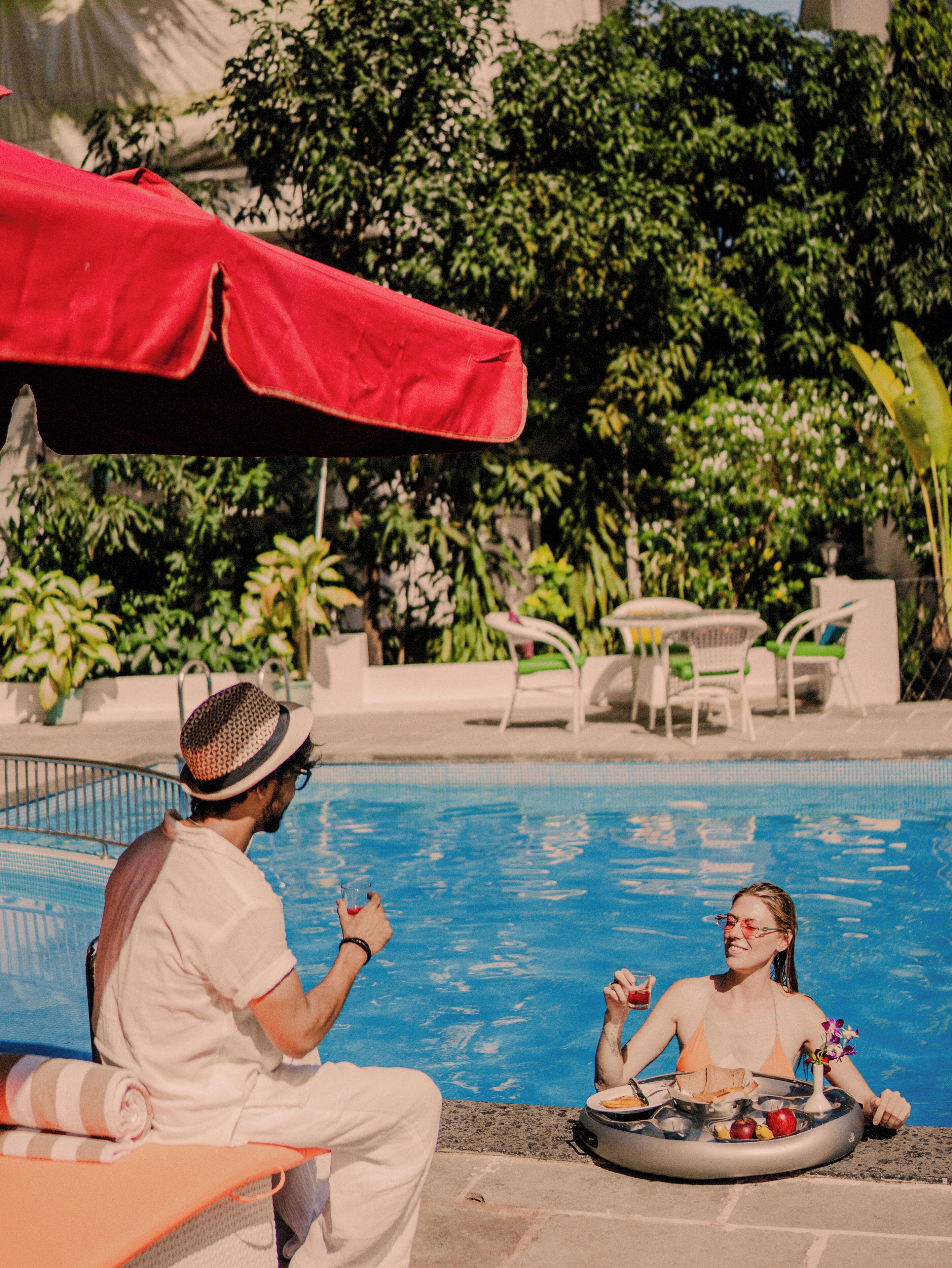Escena junto a la piscina con una sombrilla roja, sillas de descanso y mesas para comer.