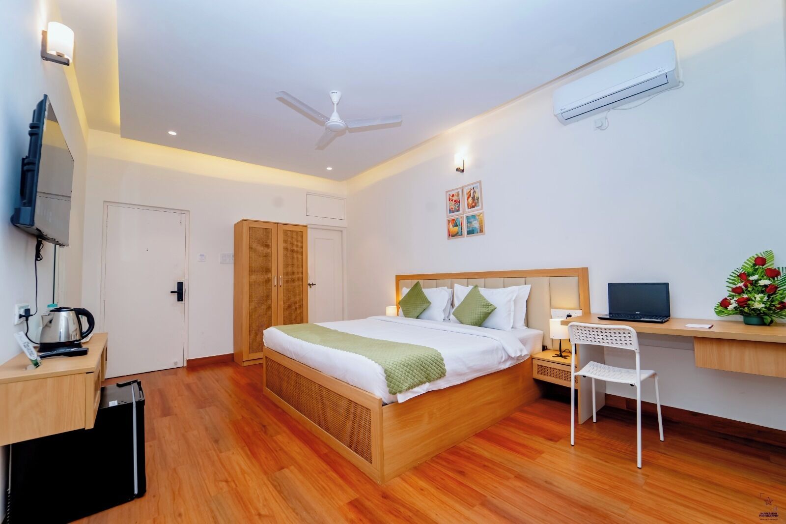 Una habitación de hotel moderna con una cama de madera, un escritorio con una computadora portátil, una silla blanca, un aire acondicionado y un televisor.
