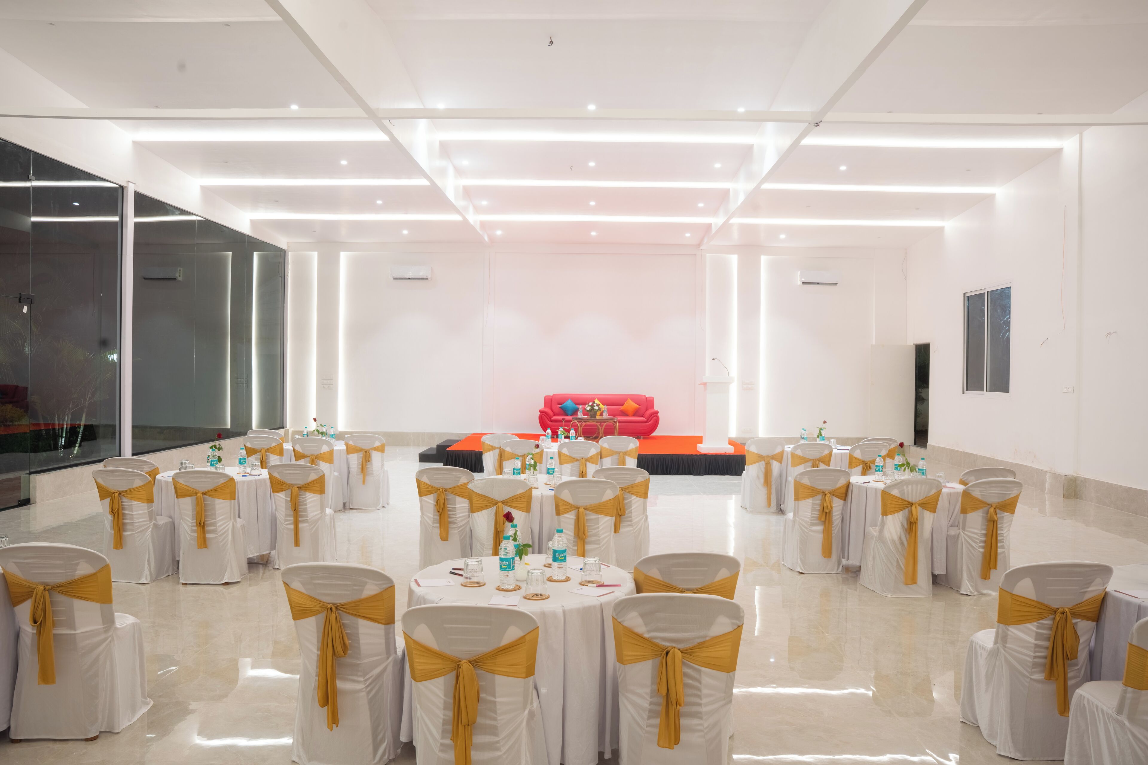 A spacious hall with round tables, each surrounded by chairs with golden sashes, and a stage with a red backdrop in the background.