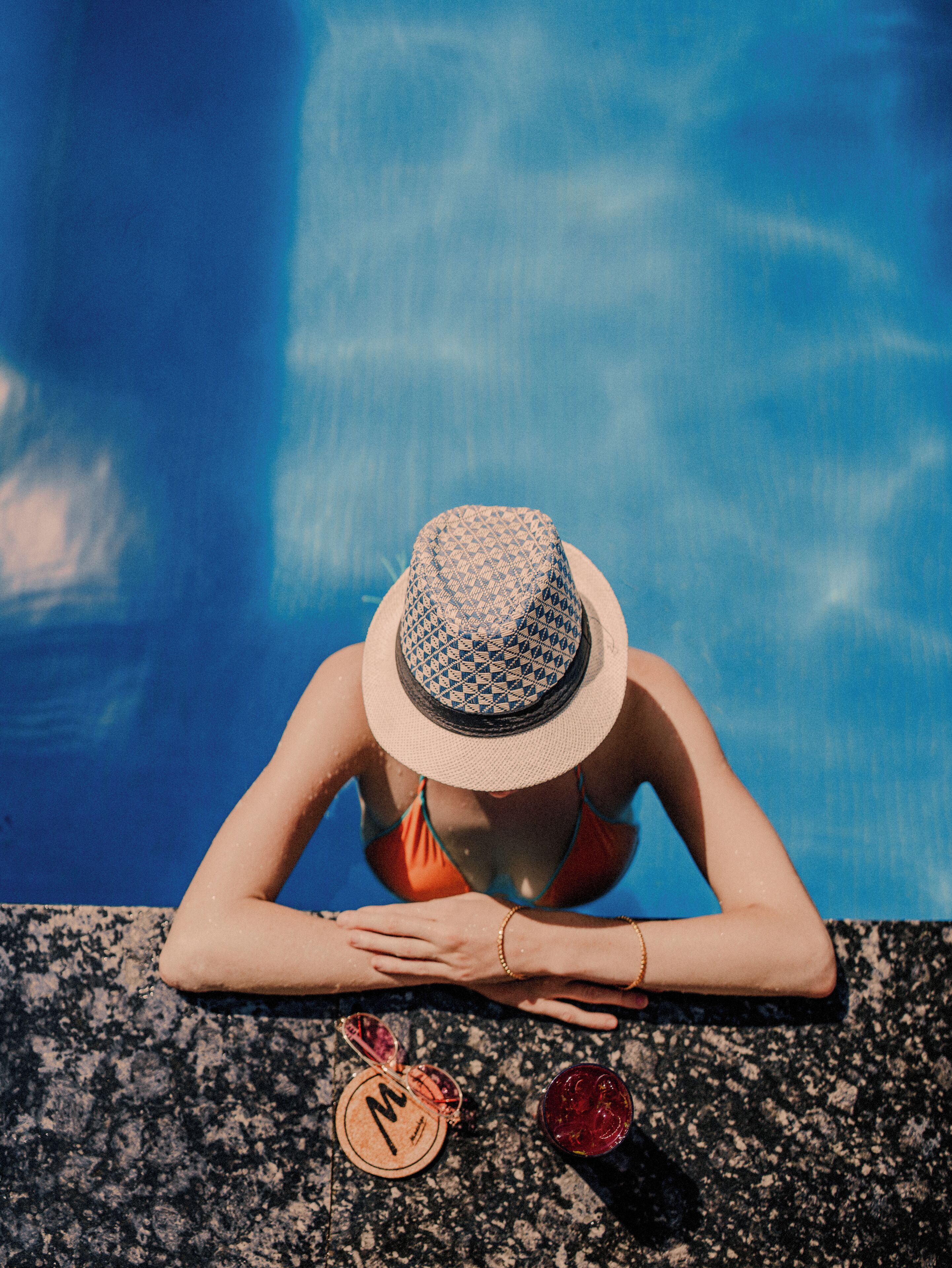 A person leaning on a pool edge with a hat, sunglasses, and a drink on a coaster.