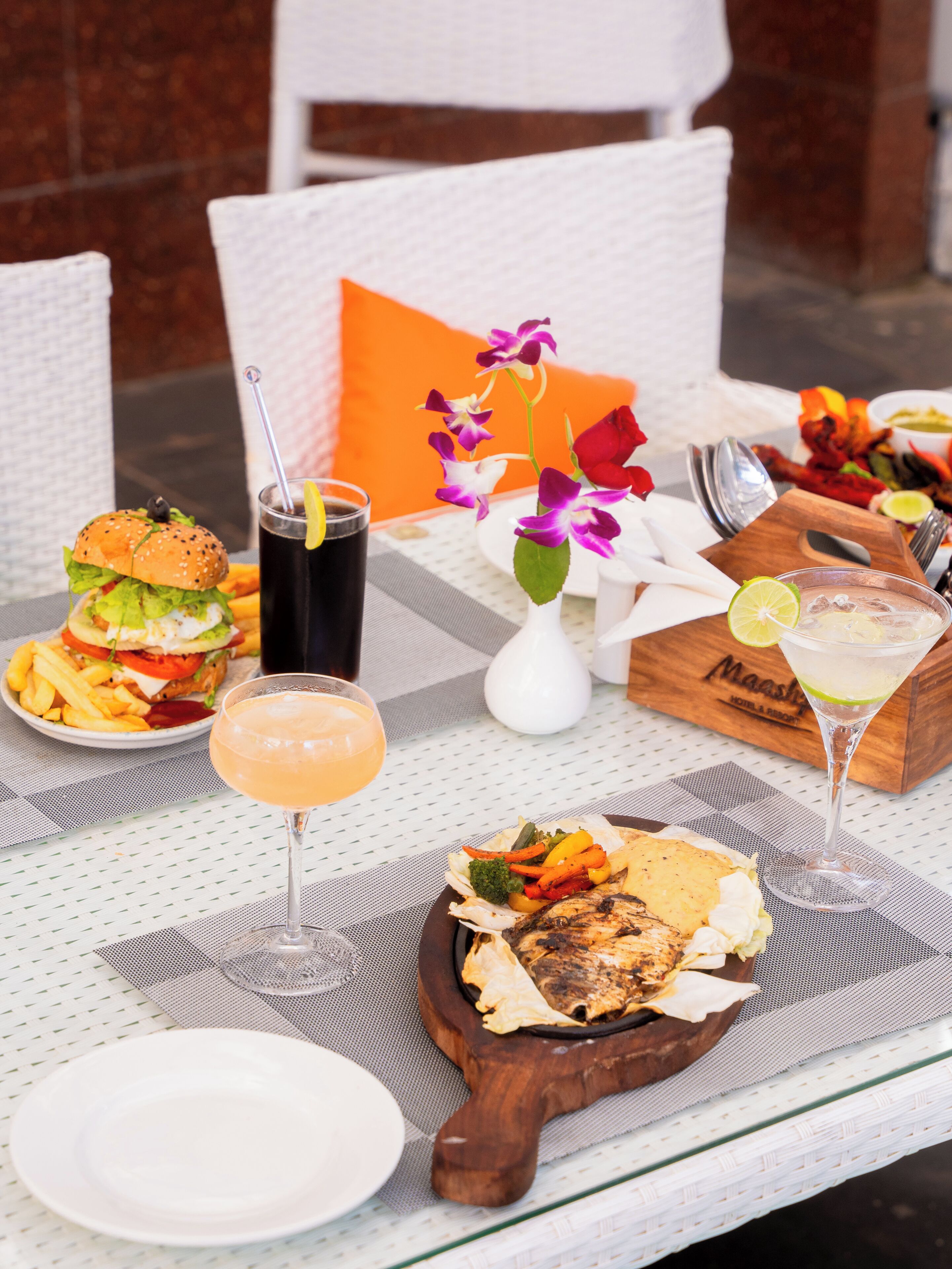 Une table dressée avec un hamburger, des frites, un cocktail margarita et un plat grillé, agrémentée d’un vase de fleurs et d’une boîte en bois portant l’inscription « Maasai Hotel & Resort ».