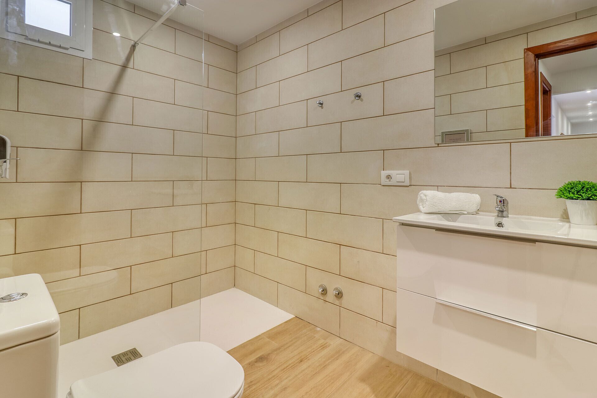 Un baño moderno con azulejos color beige, un inodoro blanco, un lavabo con espejo y una pequeña planta en maceta.
