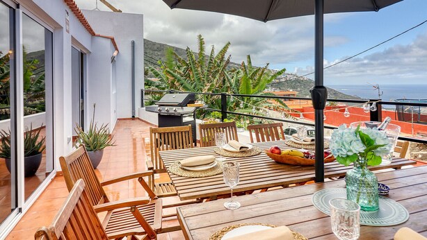 An outdoor patio with wooden furniture, a grill, and a view of the sea and hills. - Cozy House w/ BBQ, Teide Views, Near Town (Icod de los Vinos)