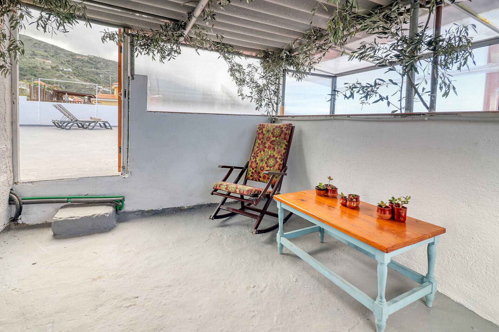 Un patio techado con una mecedora, una mesa de madera y plantas en macetas.