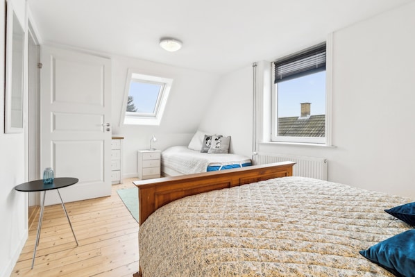 Un dormitorio con una cama de madera, una mesita de noche pequeña, un buró y una ventana con persianas, además de un tragaluz.