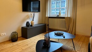 A modern living room with a flat-screen TV mounted on the wall, a large window with curtains, a dark-colored console table with decorative items, and a round coffee table with a bowl and a smartphone. - Tante Hedwig - cozy vacation apartment in Rodalben close to the rock hiking trail (Rodalben)