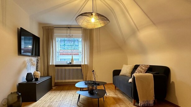 A cozy living room with a wooden floor, a black sofa with a throw blanket, a coffee table, a TV on the wall, and a large window with curtains. - Tante Hedwig - cozy vacation apartment in Rodalben close to the rock hiking trail (Rodalben)