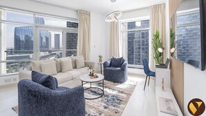 A modern living room with a sofa, two armchairs, a coffee table, and a desk with a laptop. Large windows offer views of the cityscape.