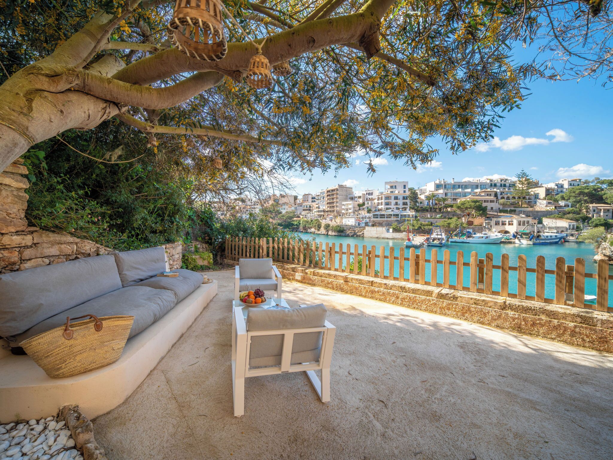 A coastal outdoor seating area with a sofa and a table, a wooden fence, and a view of a harbor with boats and buildings.