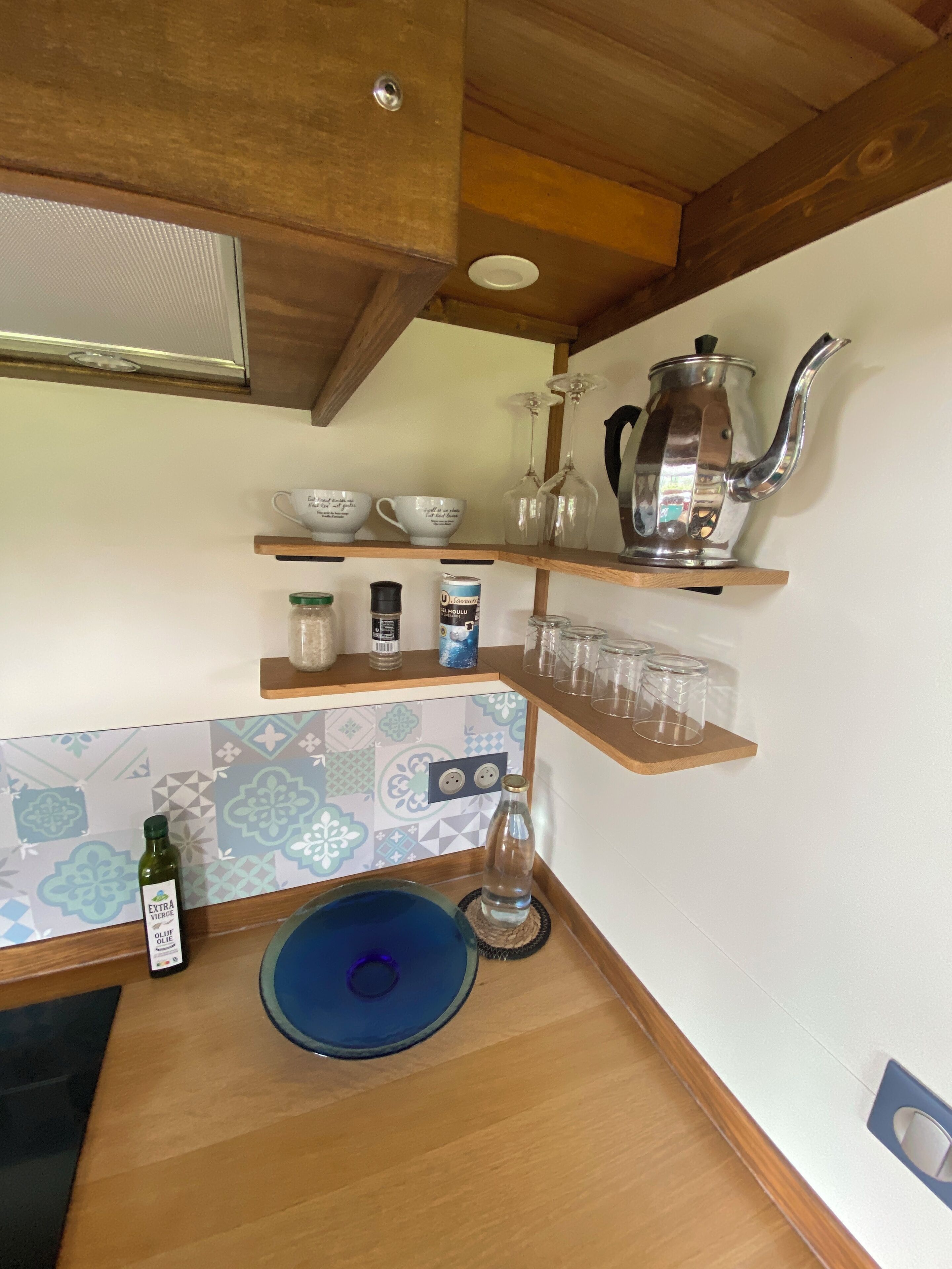 Una cucina con scaffali in legno che contengono tazze, una caffettiera e barattoli. Sul bancone si trova una bottiglia d'olio d'oliva e una ciotola blu.