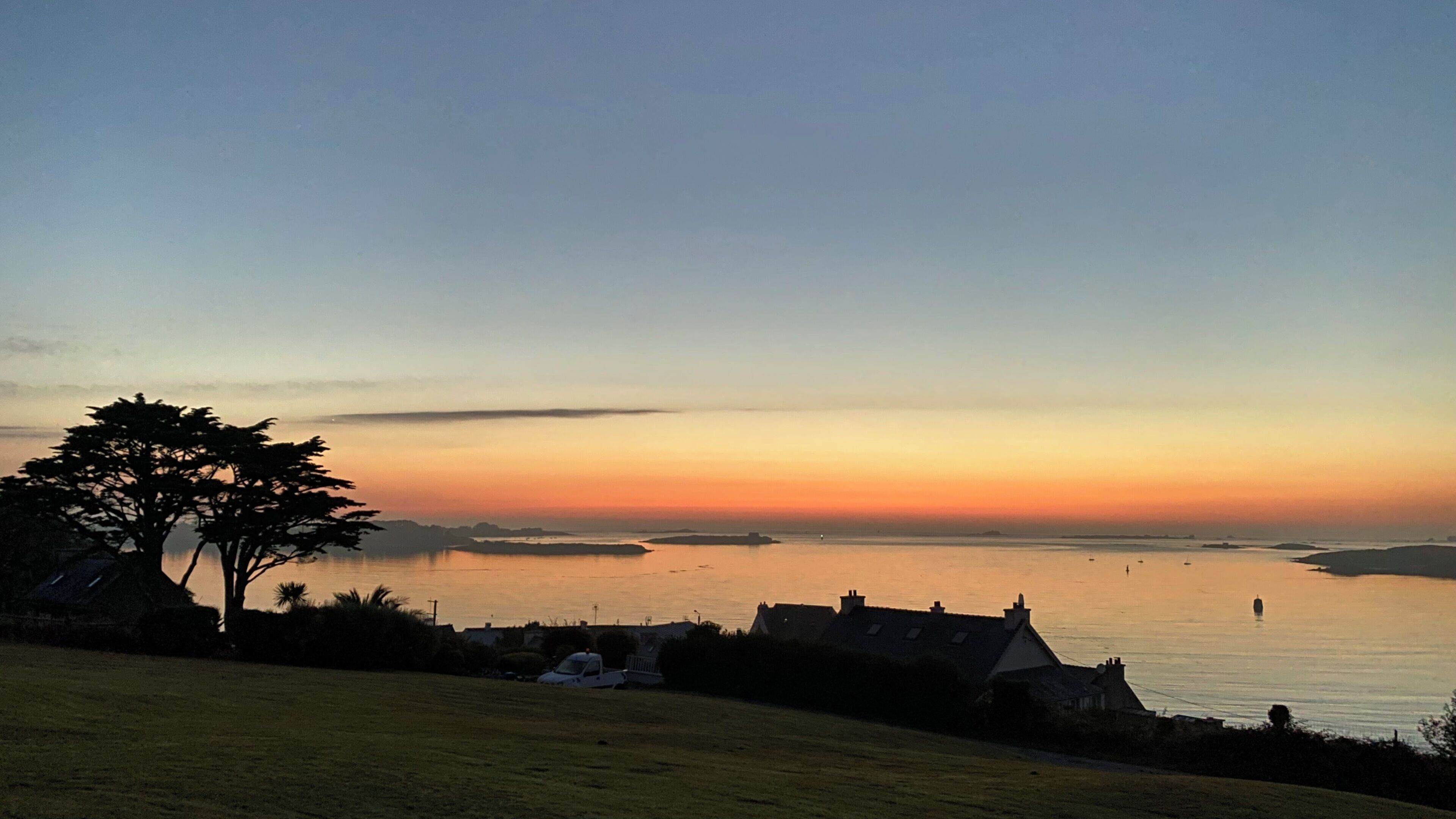 A coastal landscape with a clear sky during sunset, featuring a calm sea, a prominent tree, and a few houses in the foreground.