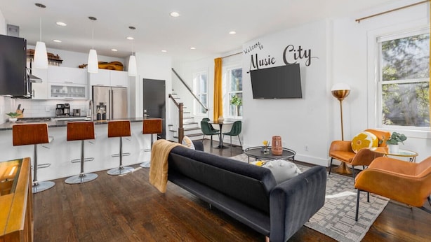 A modern living room with a black sofa, wooden bar stools, and a dining area with a table and chairs.