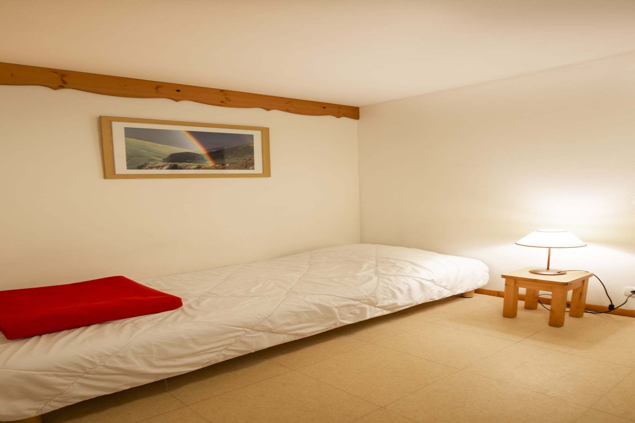 Een eenpersoonsbed met een rood kussen, een klein houten nachtkastje met een lamp, en een lijst aan de muur.