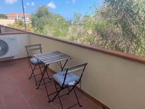 A balcony with a table and two chairs, an air conditioning unit, and a view of greenery and buildings. - Emerald Apartment. Just a few steps from the ocean (Sardegna)