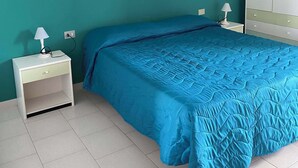 A bedroom with teal walls, a blue bedspread, a white nightstand with a lamp, and a wardrobe.