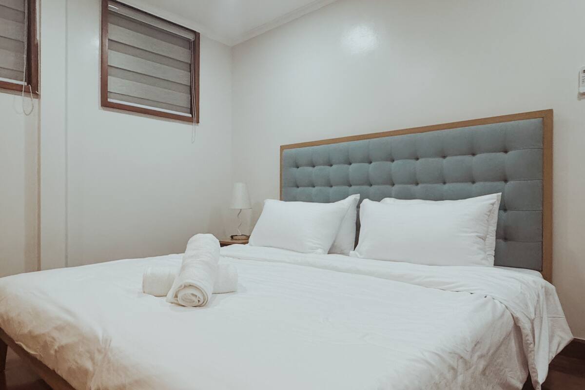 A neatly made bed with white linens and a rolled towel, a grey tufted headboard, and a bedside lamp on a wooden nightstand.
