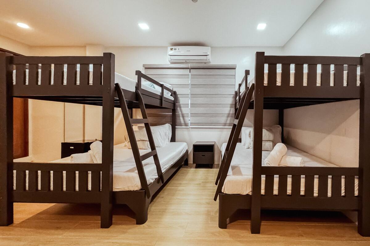 A dormitory room with bunk beds, a small desk, and a window with blinds.