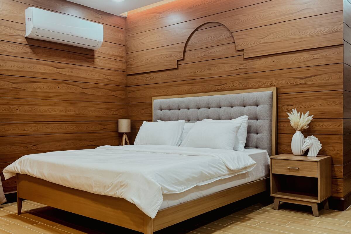 A neatly arranged bedroom with a wooden headboard, a bed with white linens, a bedside table with a lamp, and a small cabinet with decorative items.