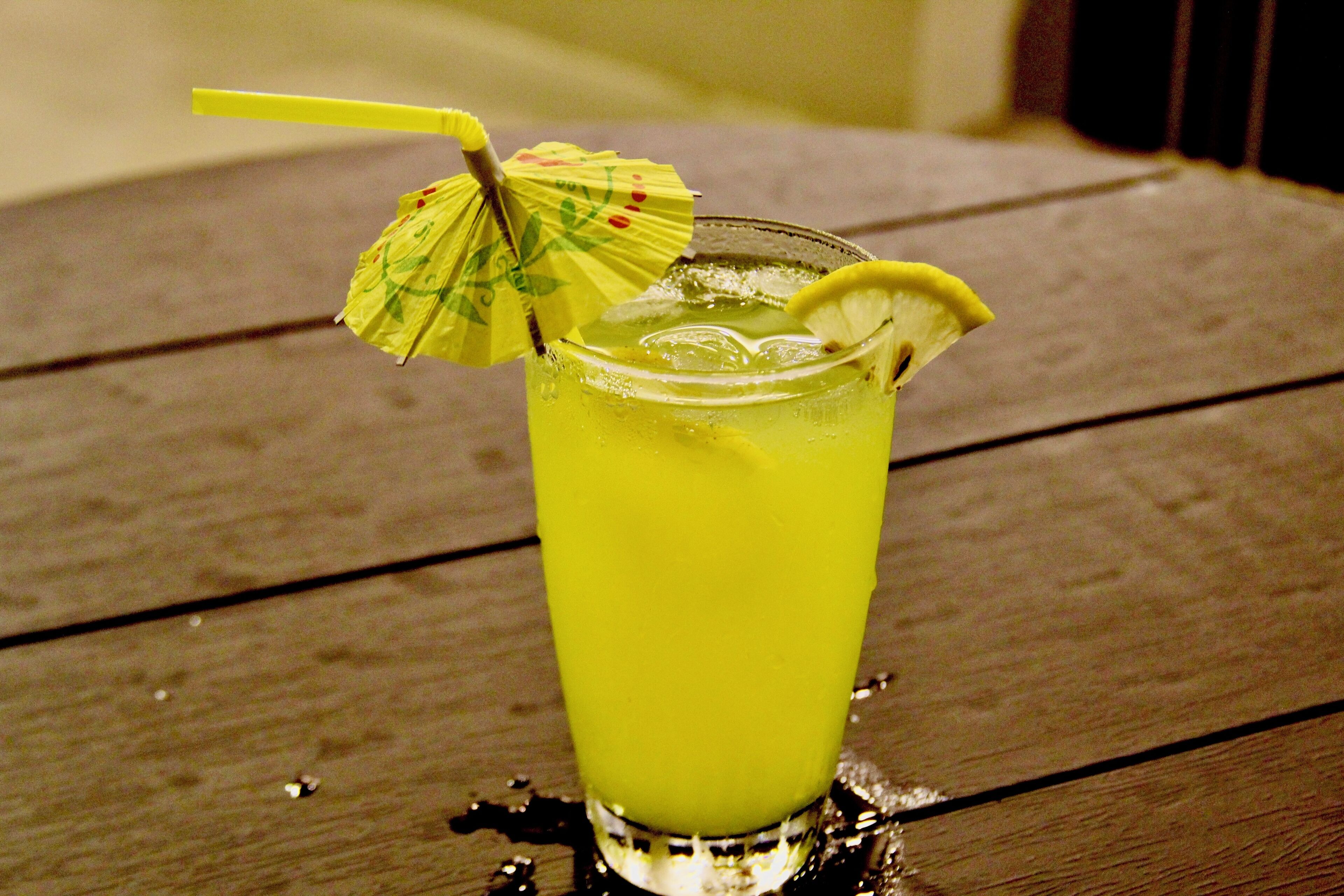 Un vaso de bebida amarilla con una rodaja de limón y un paraguas amarillo sobre una mesa de madera.