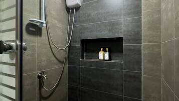Un área de ducha moderna con azulejos oscuros, un cabezal de ducha manual, un panel de control en la pared y un estante con artículos de aseo.
