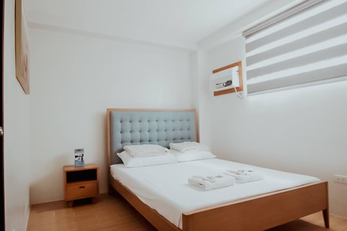 A hotel room with a wooden bed frame, a tufted headboard, a nightstand, and a window with blinds.