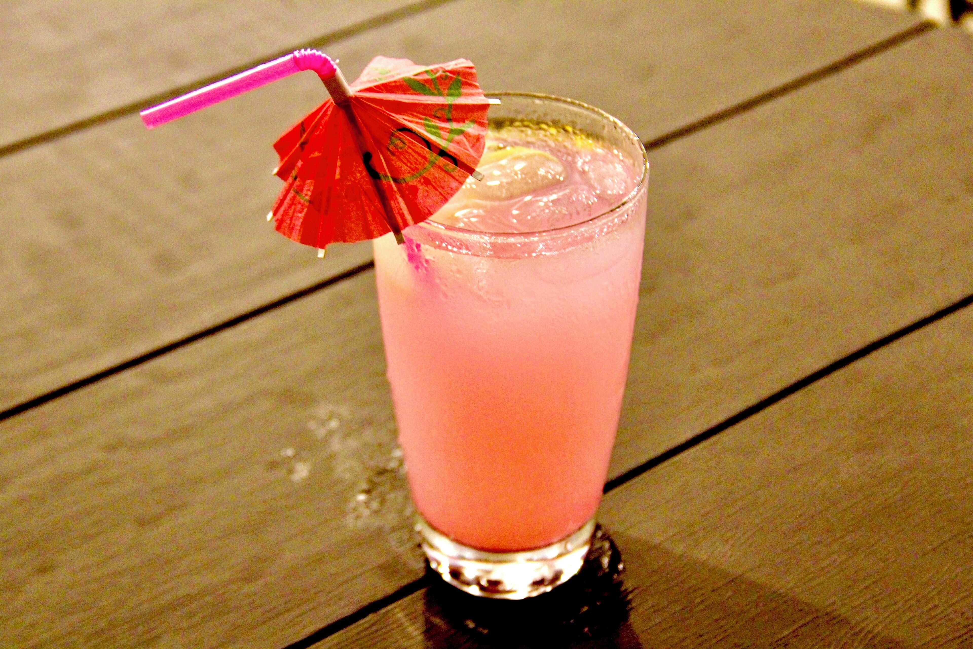 A glass of pink beverage with a straw and a decorative umbrella on a wooden table.