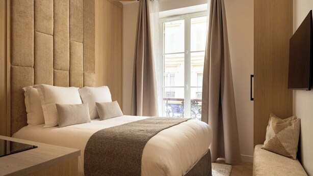 Une chambre moderne avec un grand lit, une table de chevet et une vue sur l’extérieur par une fenêtre agrémentée de rideaux.