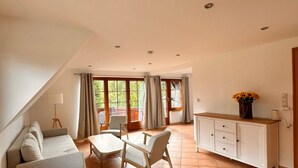 A spacious living area with a sofa, dining table, chairs, and a cabinet. Large windows allow natural light and provide views of greenery outside.