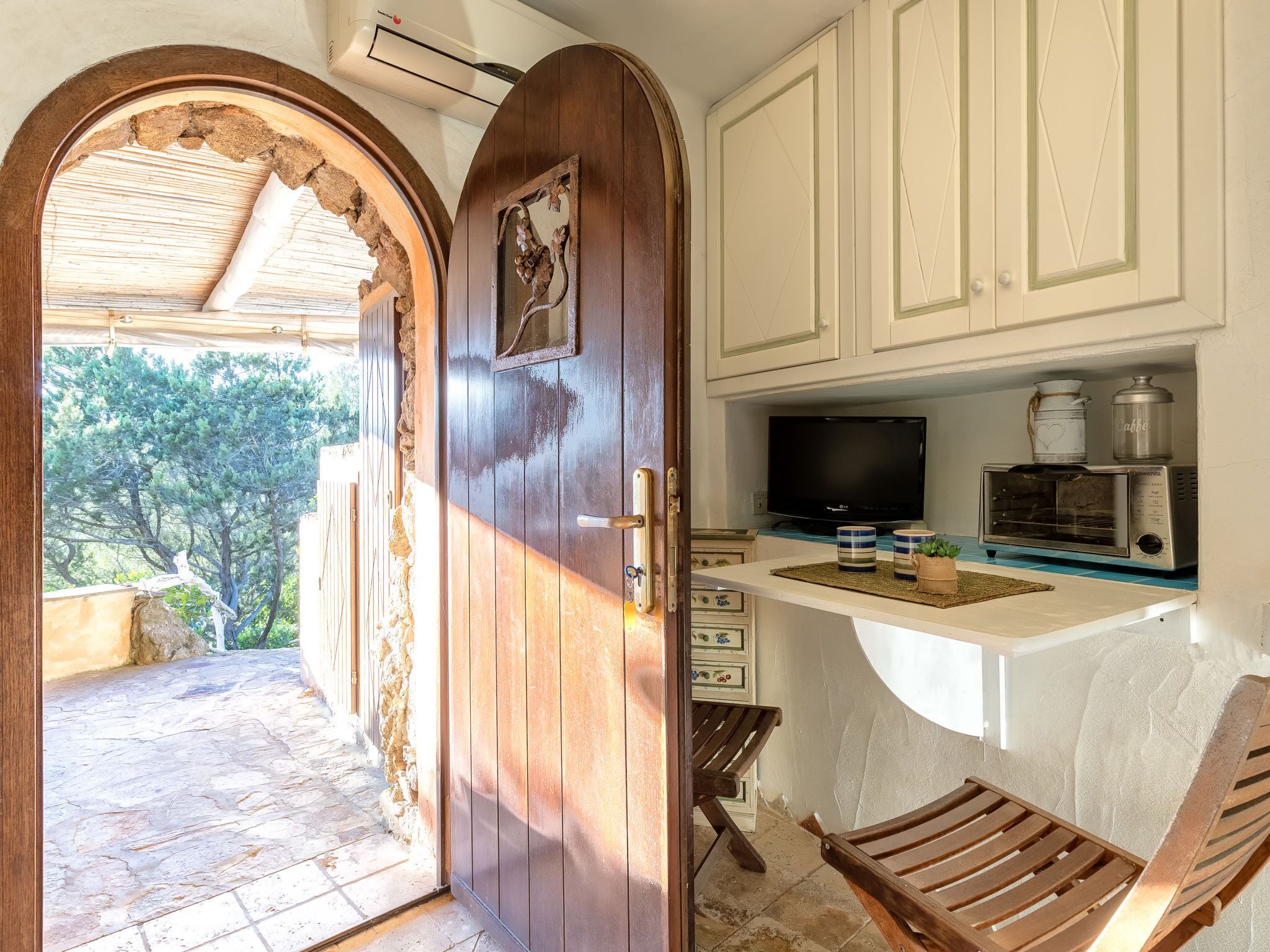 A wooden door leading to a stone-paved outdoor area with a view of trees. Inside, there is a kitchenette with a TV, a coffee maker, and a toaster oven on a white countertop.