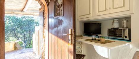 A wooden door leading to a stone-paved outdoor area with a view of trees. Inside, there is a kitchenette with a TV, a coffee maker, and a toaster oven on a white countertop.