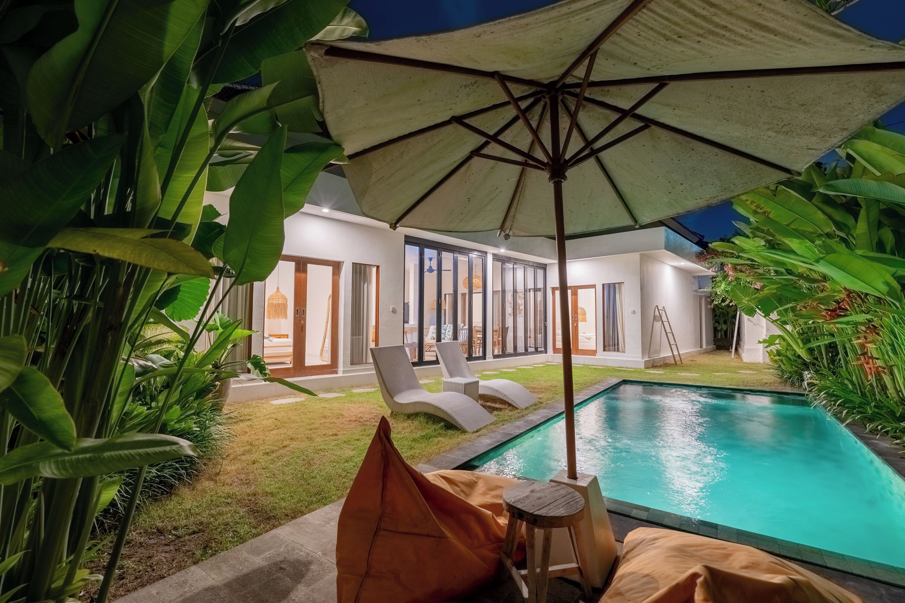 A poolside area with lounge chairs and an umbrella, surrounded by lush greenery.